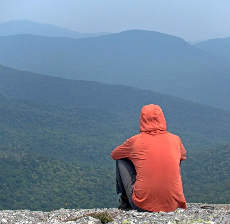 The image shows a person sitting on a rocky outcrop, gazing out at a vast landscape of rolling hills and mountains. The person is wearing a hooded orange shirt and dark pants, their back to the viewer. The hills are covered in lush greenery, fading into a hazy blue in the distance. The sky is overcast, creating a soft, diffused light. The overall scene evokes a sense of solitude, contemplation, and appreciation for nature's grandeur.